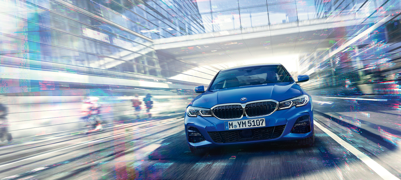 Vista frontal del BMW Serie 3 Sedán en movimiento de día bajo un edificio de cristal.