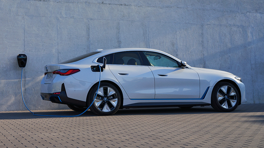 Vista del lateral con BMW Wallbox BMW i4 eDrive40 G26 2021 BMW i4 eDrive40 G26 2021 Mineralweiss Vista del lateral delante de la pared de una casa con punto de carga BMW Wallbox