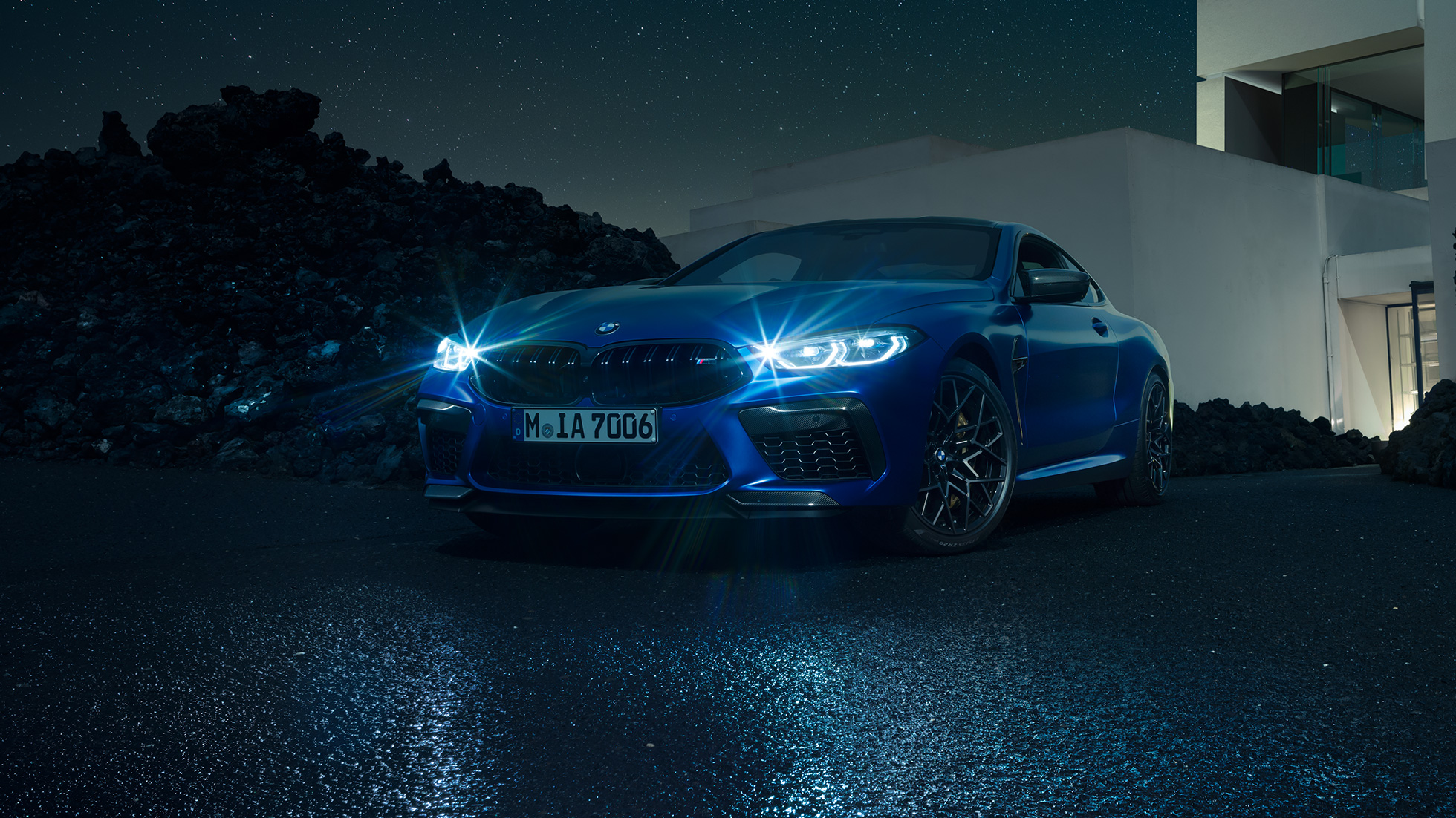 BMW M8 Competition Coupé, vista frontal tres cuartos. BMW M8 Competition Coupé en Marina Bay Blue metálico con luces LED brillantes, vista frontal tres cuartos en alto frente a una villa de noche.