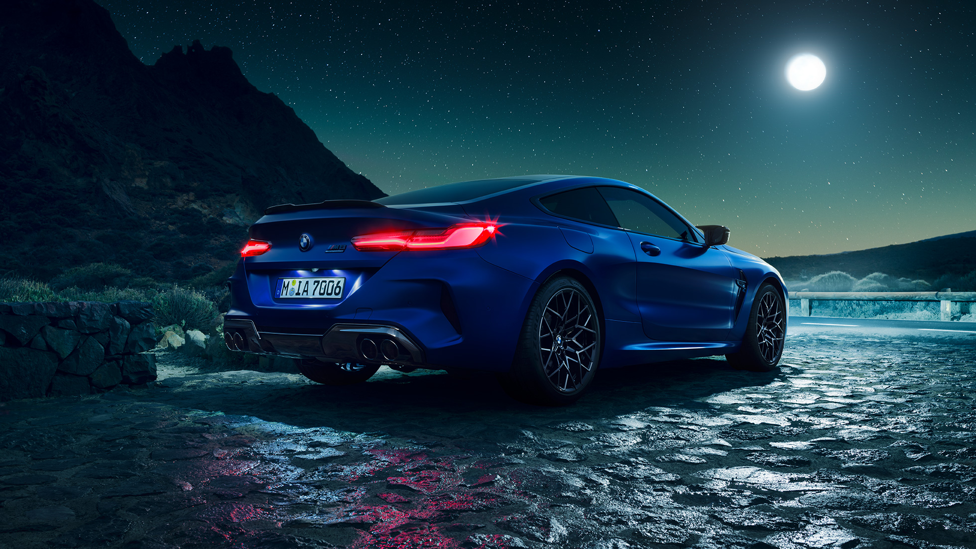 BMW M8 Competition Coupé, vista trasera tres cuartos. BMW M8 Competition Coupé en Marina Bay Blue metálico con las luces LED traseras iluminadas, vista tres cuartos en alto con luna llena.
