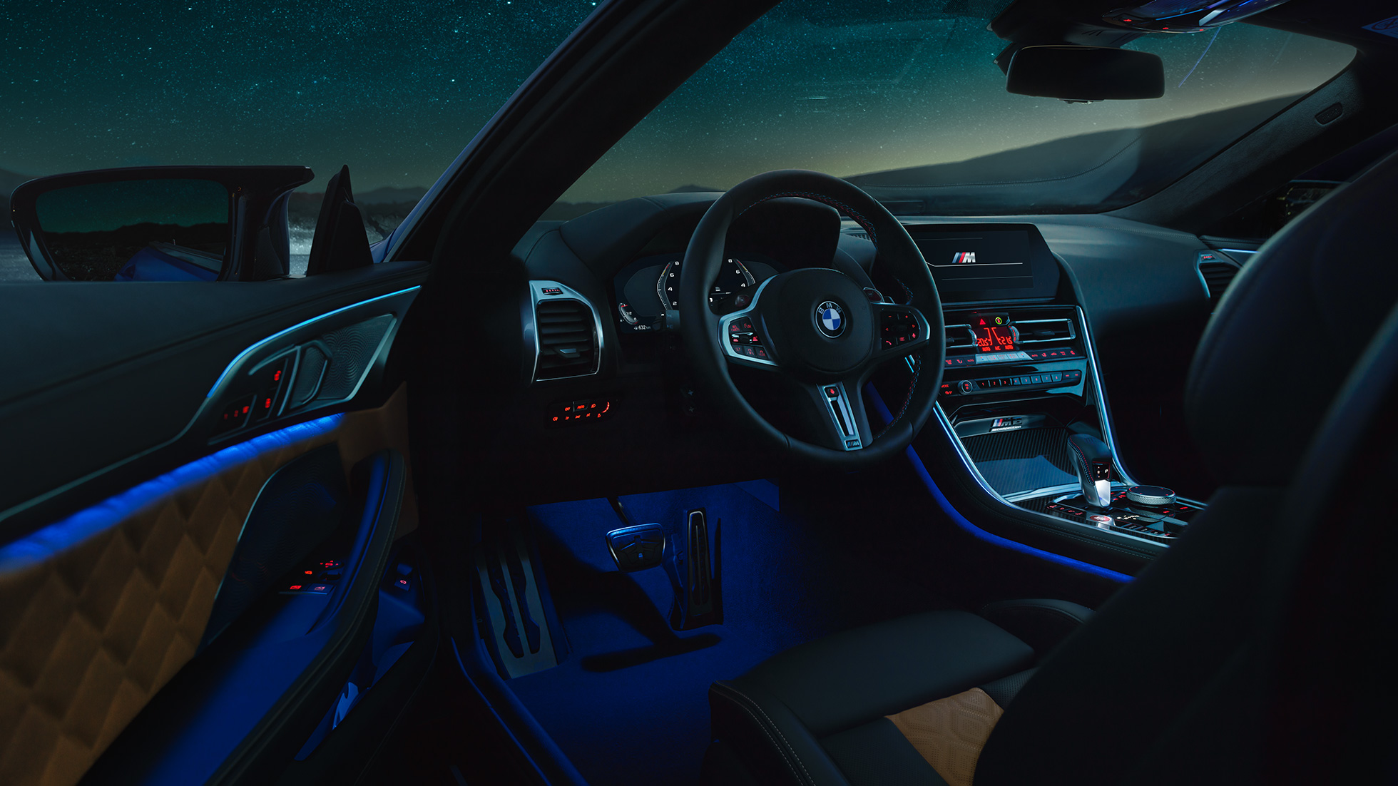 Interior BMW M8 Competition Coupé. BMW M8 Competition Coupé, interior de noche con volante en cuero M y luz ambiente azul con la puerta de conductor abierta.