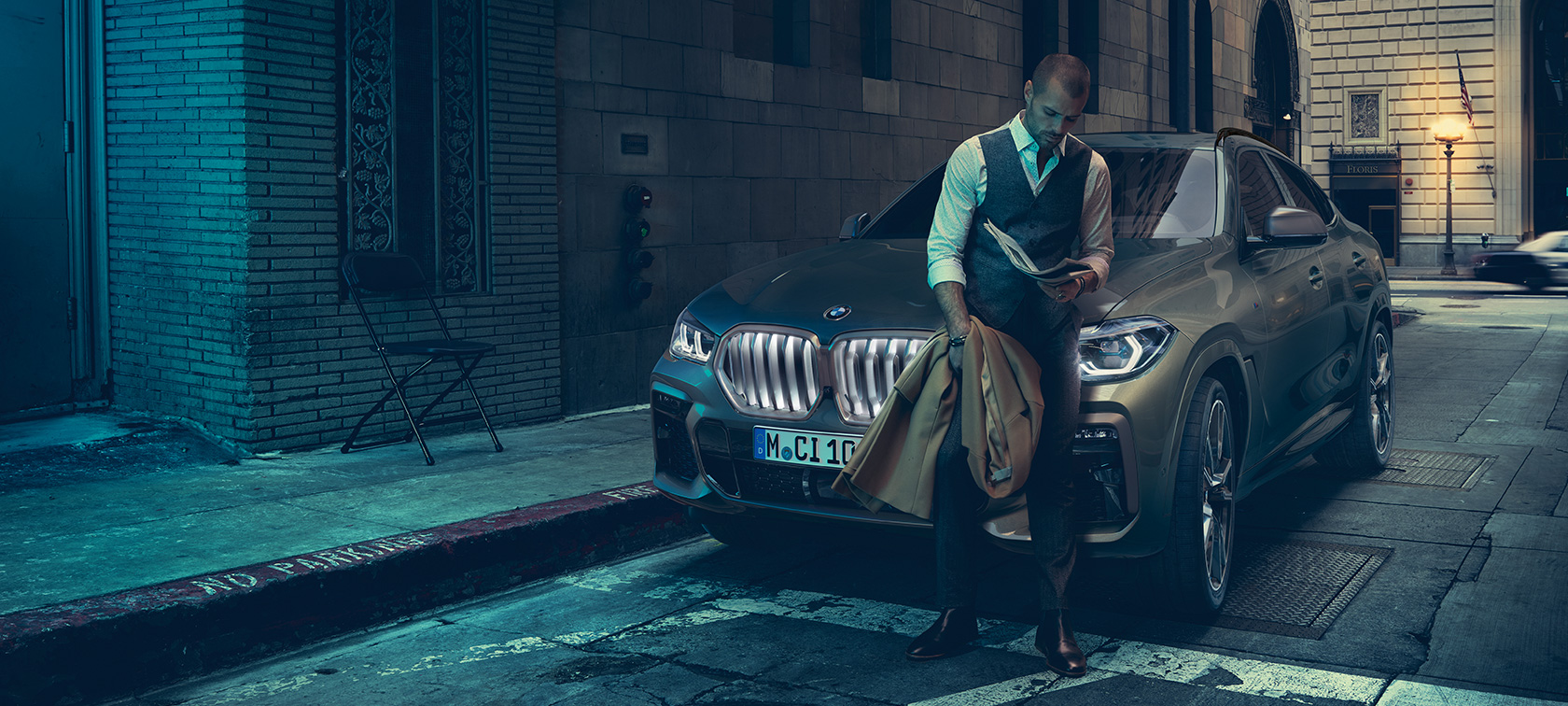 Hombre apoyado en un BMW X6. Hombre apoyado en un BMW X6 en perspectiva lateral frente a un escenario urbano.